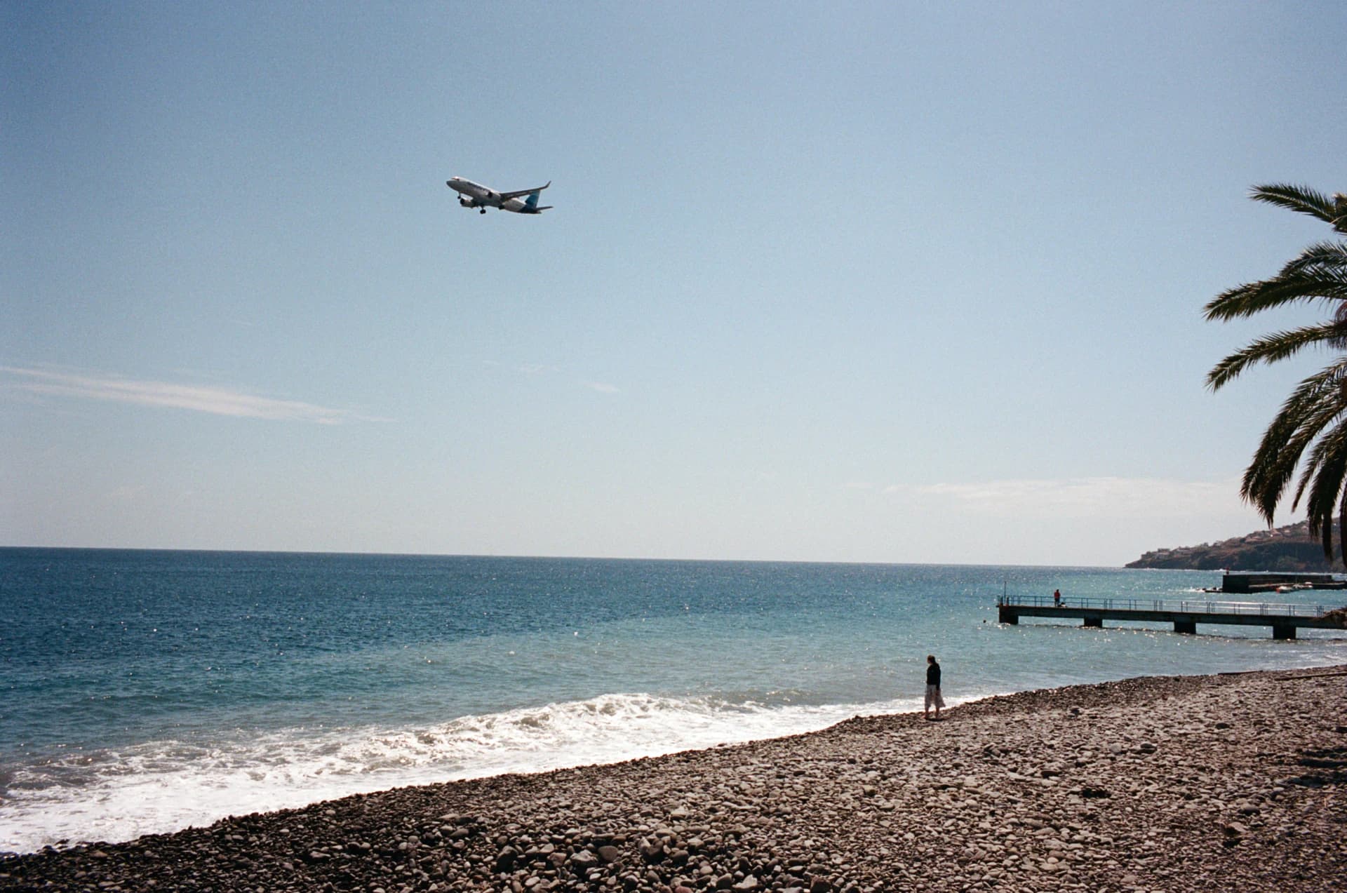 Color film photograph of a rocky shoreline with a low-flying airplane, a lone figure, and a palm tree at the edge of the frame